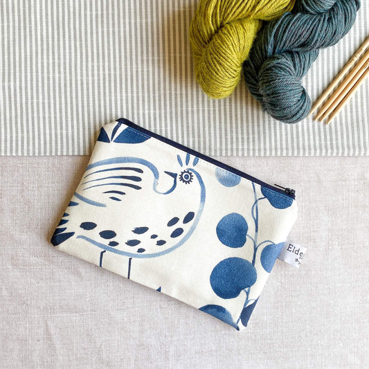 A small pouch made out of a vibrant indigo coloured folk art print lies on a table next to two skeins of yarn and some wooden knitting needles. 