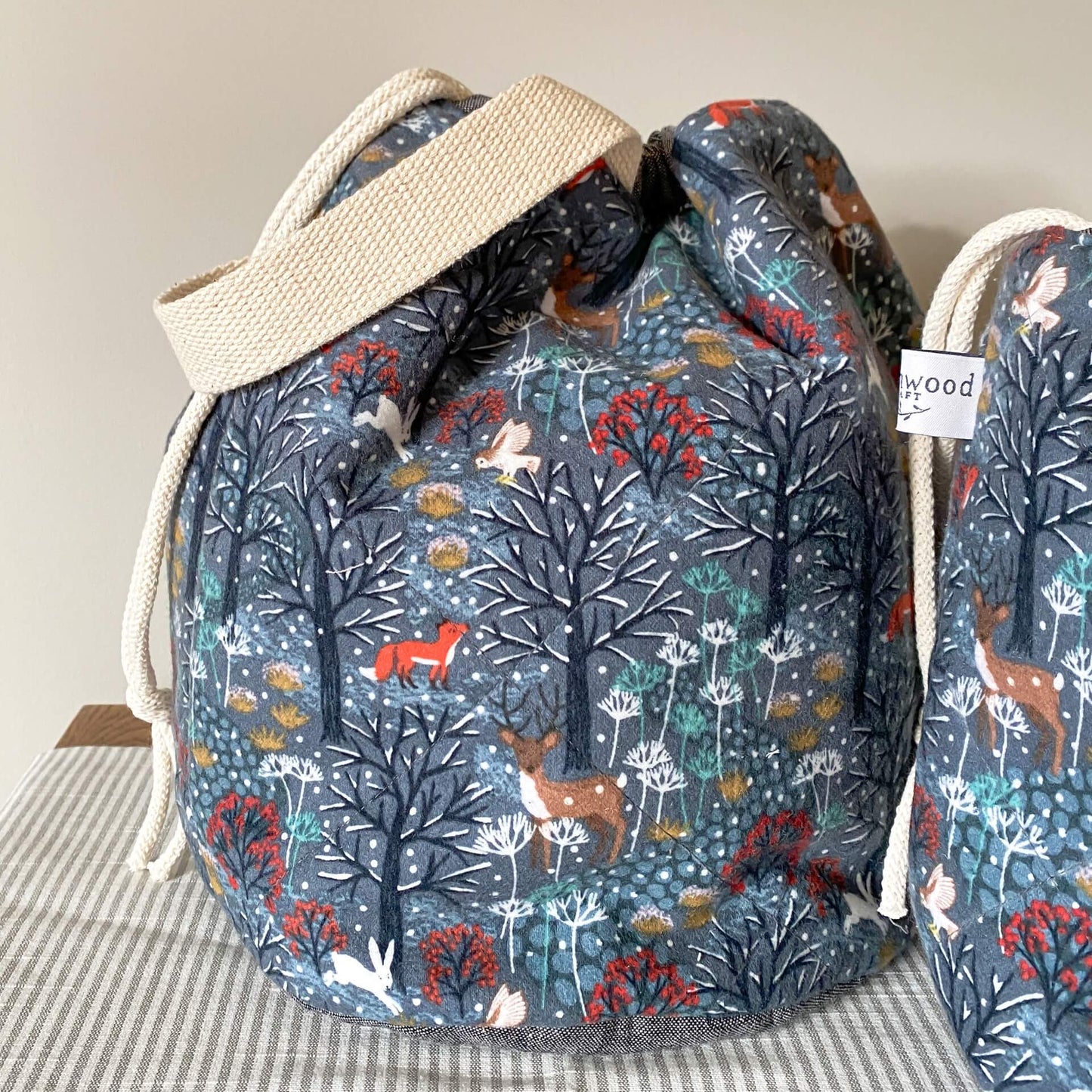 A close up picture of a project bag hand made using a winter woodland scene fabric. The fabric has a wintery grey background and shows off snowy trees, owls, rabbits, deer and foxes. The bag has a handle and drawstrings. 
