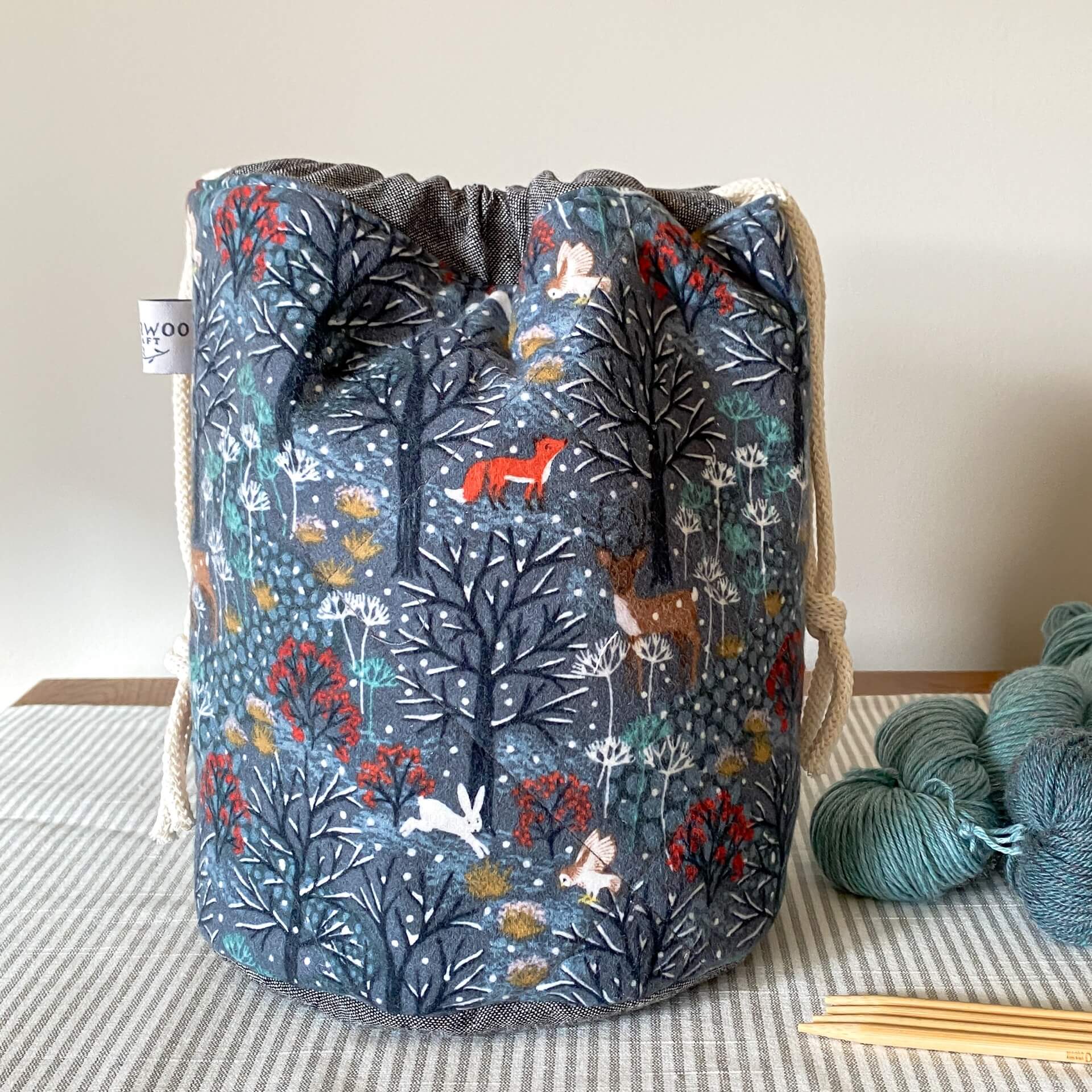 A front on view of a small project bag designed for sock knitters. This bag is made from a fabric that features a wintery woodland scene. 