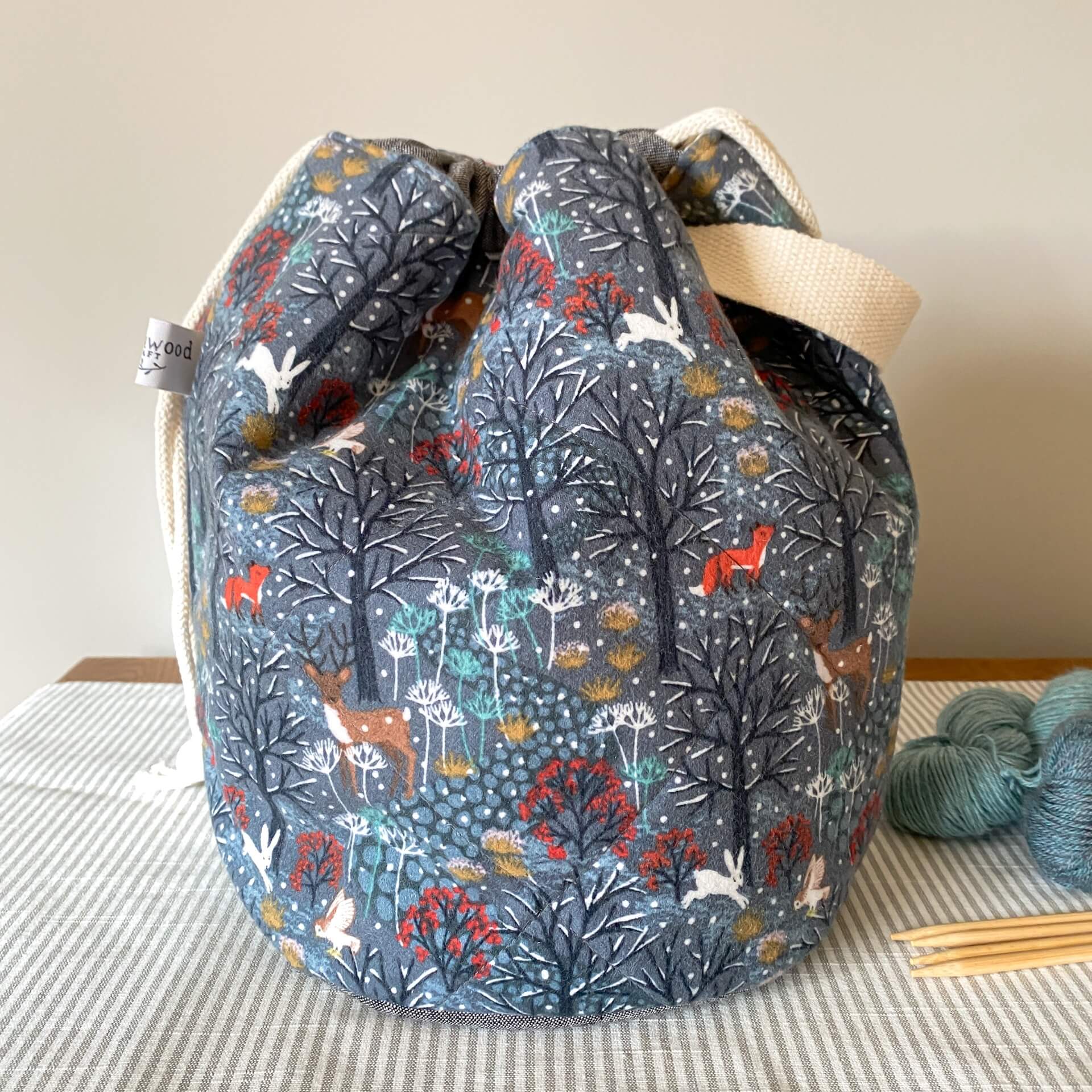A large project bag with a handle sits on a bench and is pulled closed by drawstrings. The fabric that the bag is made of shows a wintery woodland scene. 