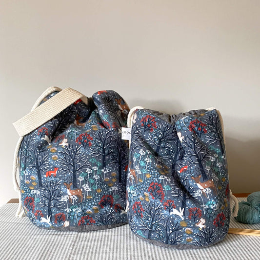 Two hand made project bags for crafters sit next to each other on a bench. The fabric the bags are made from shows a winter scene with snow tipped trees and woodland animals. 
