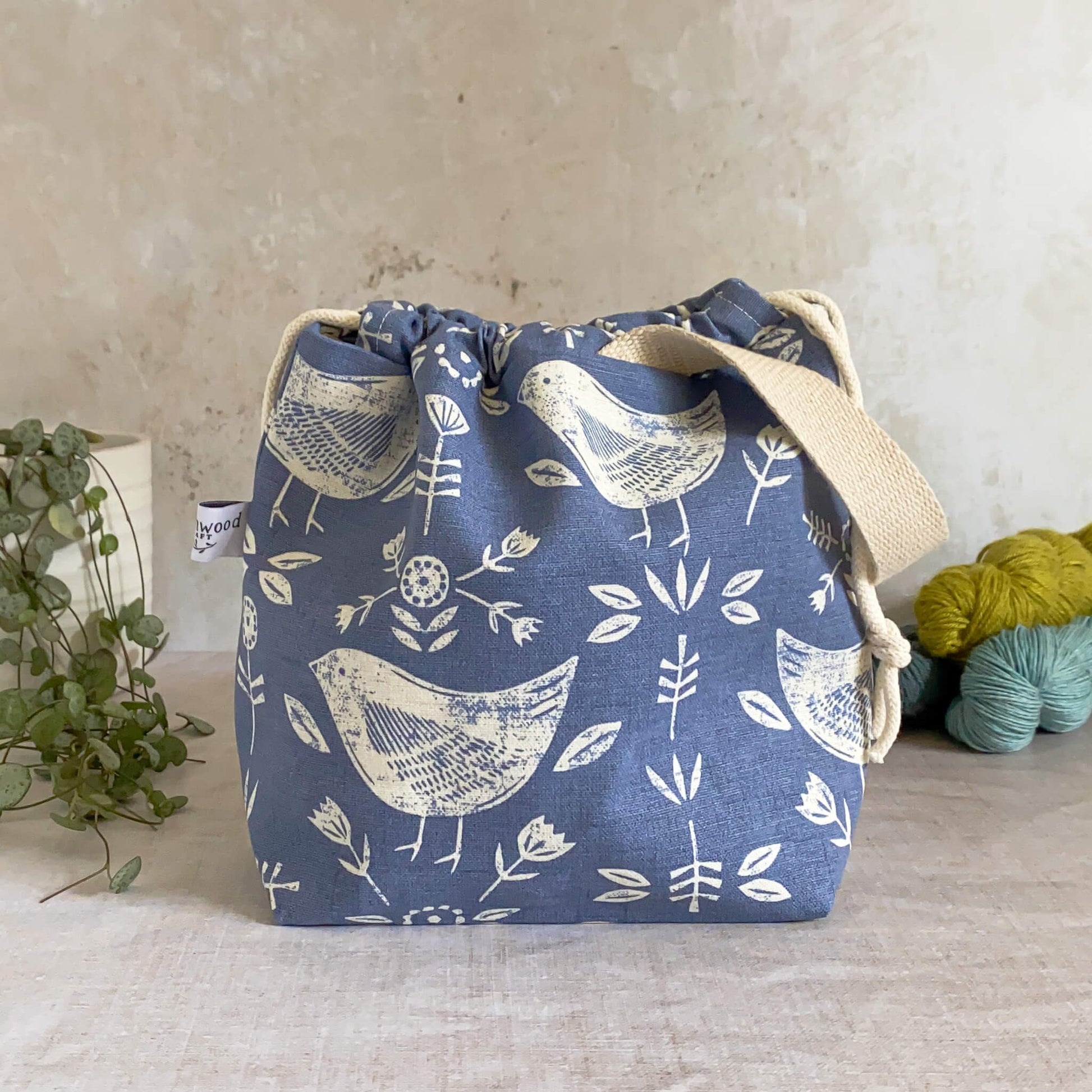 Compact handmade project bag in Scandinavian-inspired blue bird-themed fabric, equipped with a drawstring closure and sturdy handle, ideal for organizing knitting and crochet essentials. Shown alongside three skeins of yarn and a potted plant.