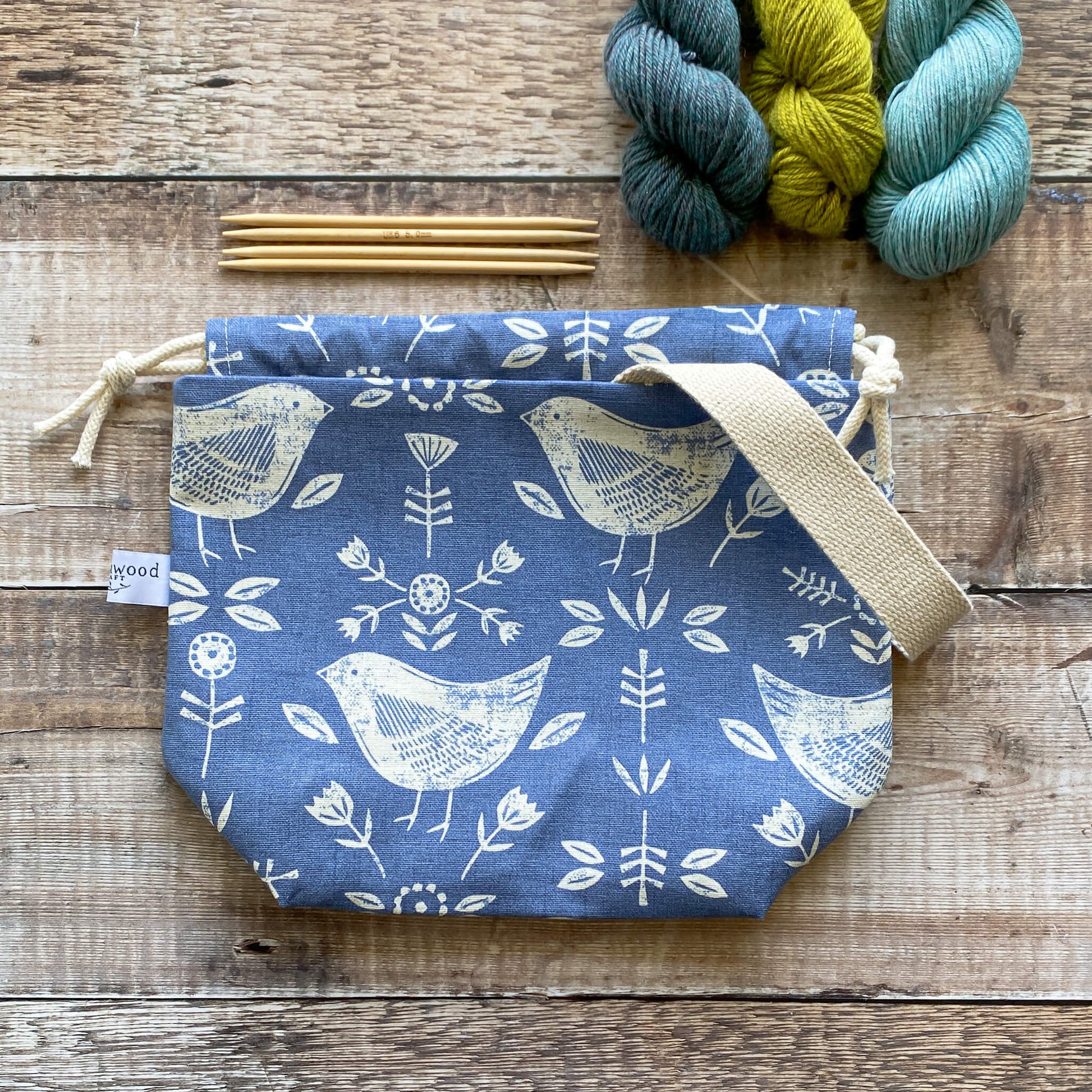 A small handmade project bag in blue bird themed fabric, designed with a drawstring closure and sturdy handle, ideal for carrying knitting or crochet supplies. The bag is displayed next to three skeins of yarn and some wooden knitting needles.