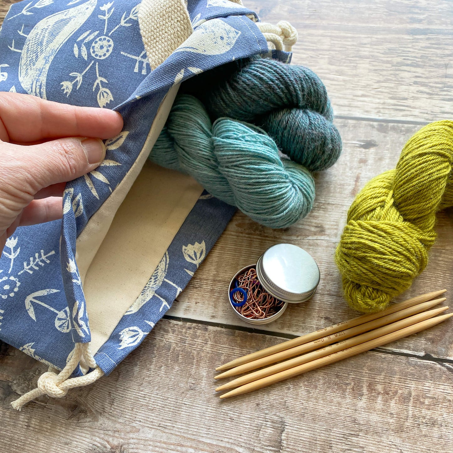 A view of the inside of a small handmade project bag for knitting and crochet projects. A pale-skinned hand holds the bag open to reveal its cream-colored canvas lining. Inside the bag, two skeins of yarn are neatly stored. Nearby, more skeins of yarn and knitting accessories are displayed. The bag is made from Scandi inspired blue bird themed fabric.