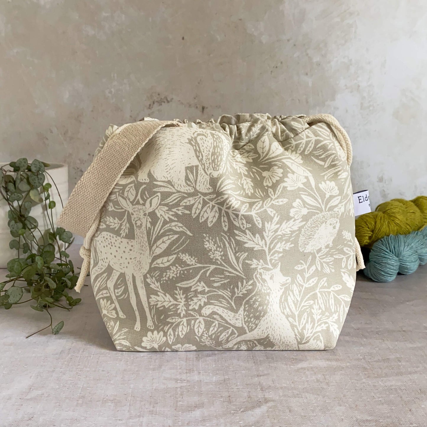 Small handmade project bag with countryside fabric, featuring a drawstring closure and sturdy handle, perfect for knitting or crochet supplies. Displayed with three skeins of yarn and a pot plant in the background.