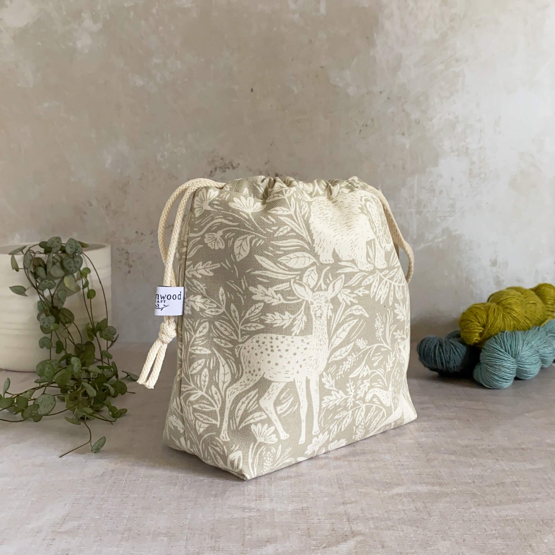 A side view of a small handmade project bag in a linen coloured countryside fabric, featuring a drawstring closure and sturdy handle, perfect for carrying knitting or crochet supplies. The bag is displayed in front of three skeins of yarn and a pot plant