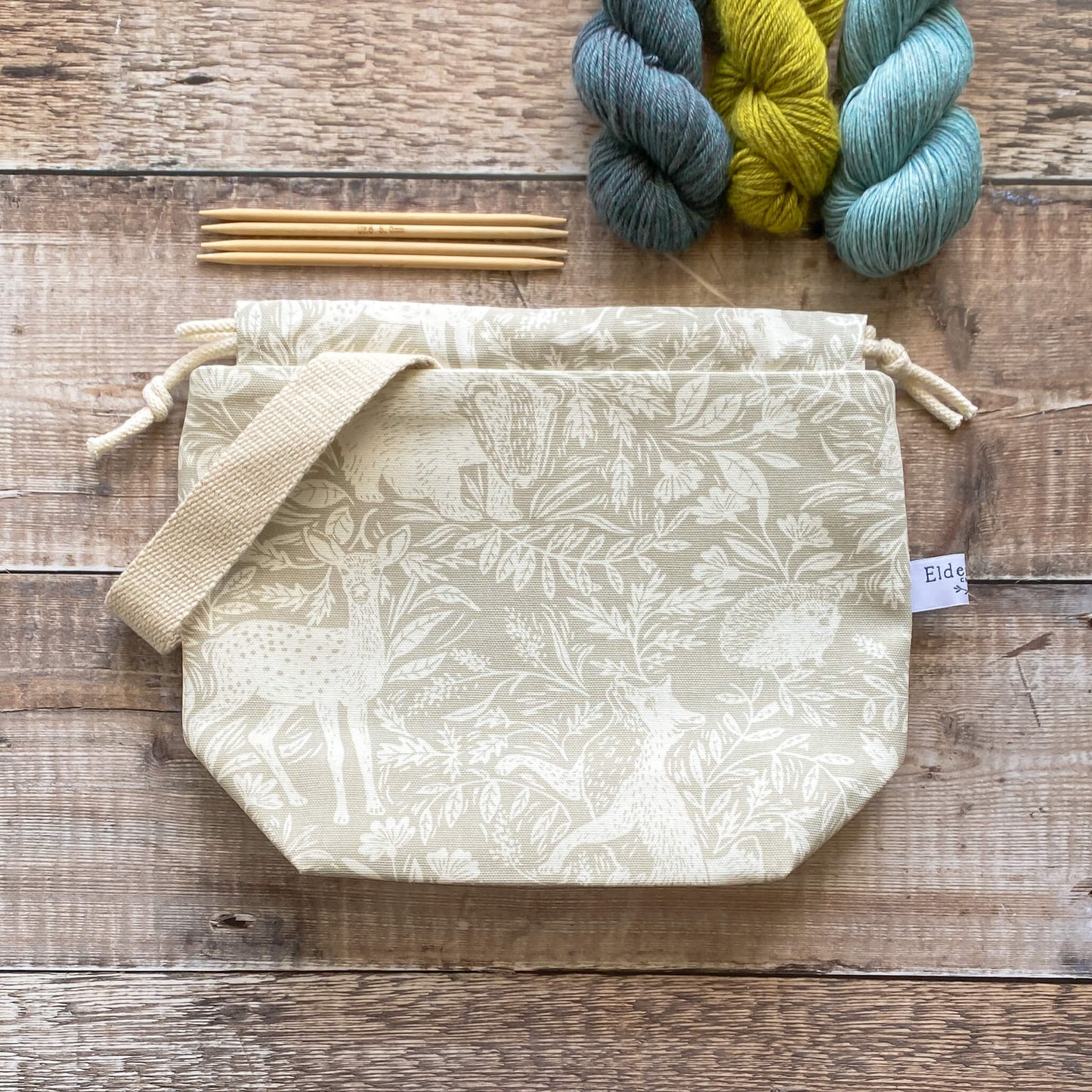 A small handmade project bag in a linen coloured countryside fabric, featuring a drawstring closure and sturdy handle, perfect for carrying knitting or crochet supplies. The bag is displayed next to three skeins of yarn and some wooden knitting needles. 