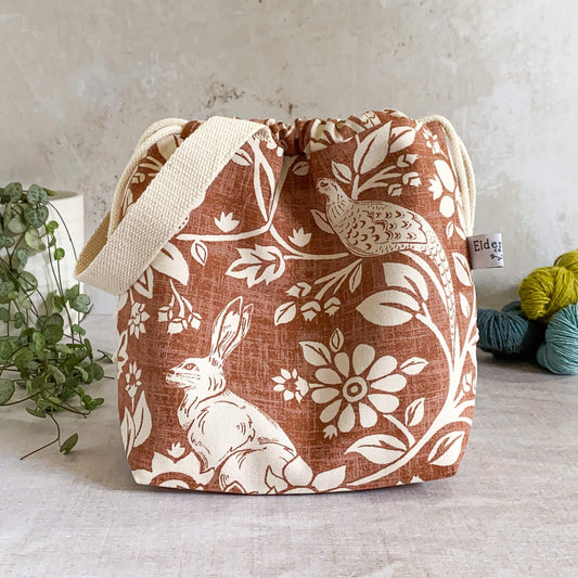 A small sized handmade project bag in a copper coloured heathland fabric. A drawstring closure at the top, and a sturdy handle, ideal for organising knitting and crochet supplies and projects. The project bag sits in front of three skeins of yarn and a pot plant. 