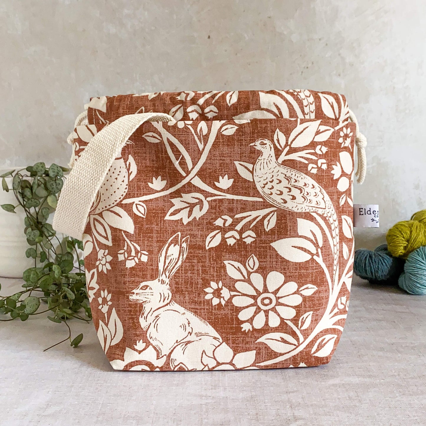 Small handmade project bag in copper-colored heathland fabric, featuring a drawstring closure and sturdy handle, perfect for organizing knitting and crochet supplies. Displayed with three skeins of yarn and a pot plant in the background.