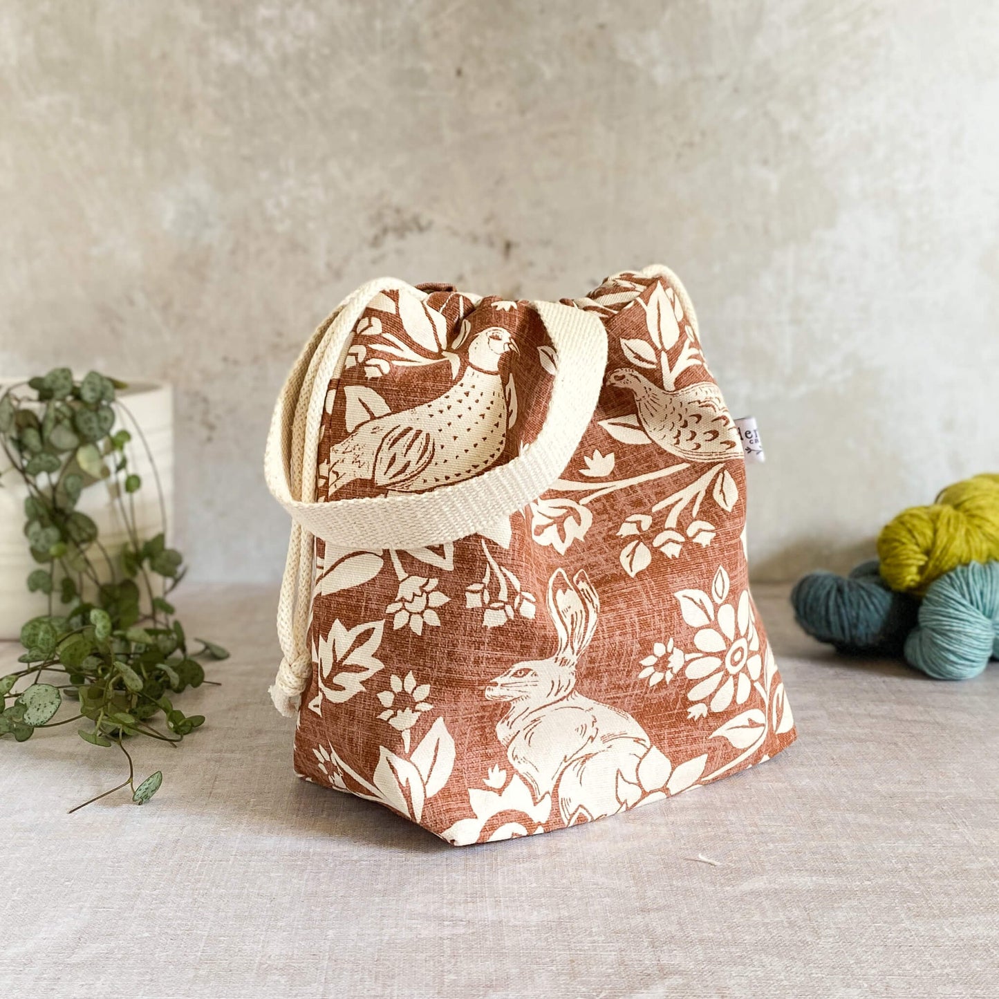 A side on view of a small sized handmade project bag in a copper coloured heathland fabric. A drawstring closure at the top, and a sturdy handle, ideal for organising knitting and crochet supplies and projects. The project bag sits in front of three skeins of yarn and a pot plant. 