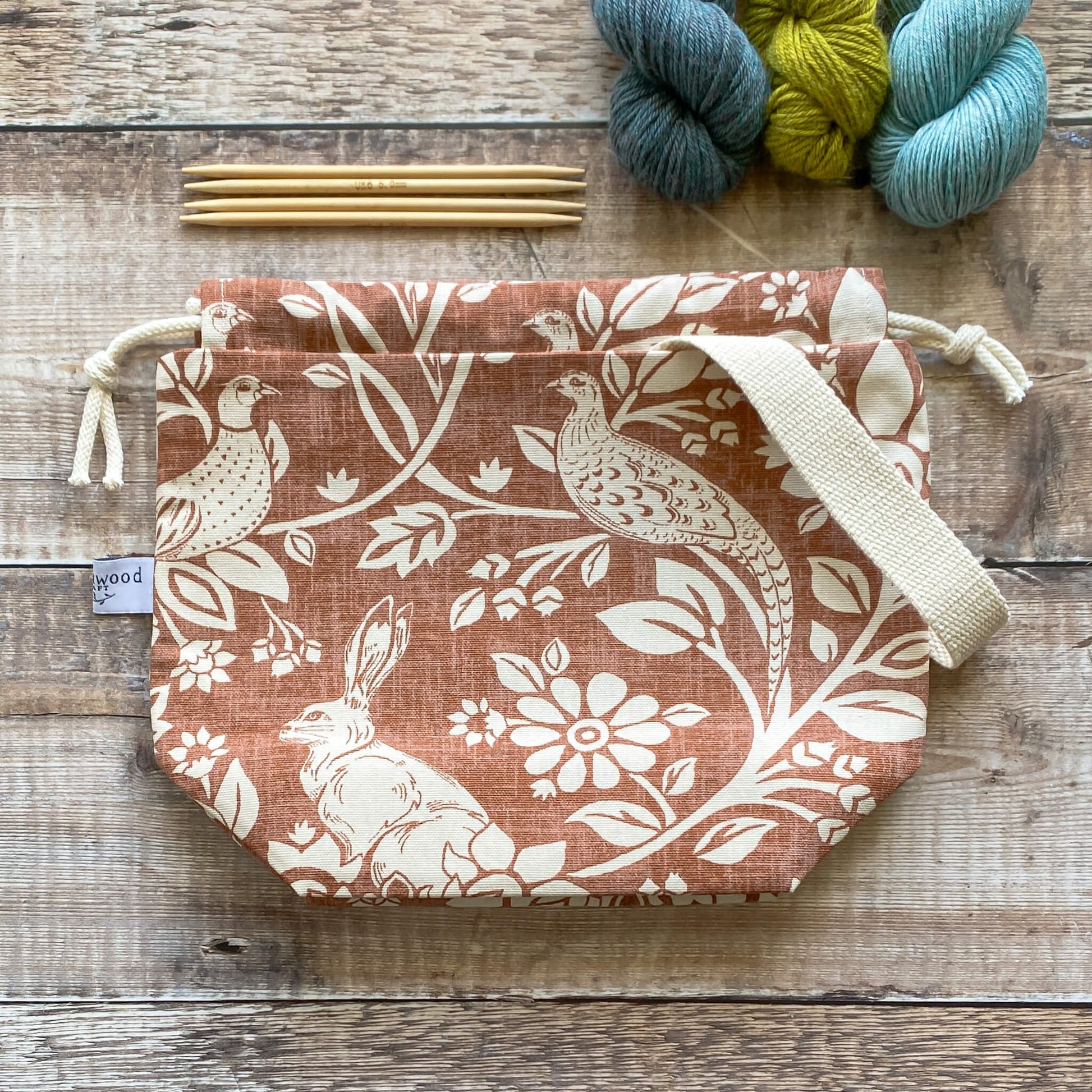 A top down view of a small sized handmade project bag in a copper coloured heathland fabric. A drawstring closure at the top, and a sturdy handle, ideal for organising knitting and crochet projects. The project bag sits next to three skeins of yarn and four wooden knitting needles.  