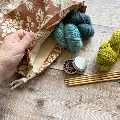 A view of the inside of a small sized handmade project bag for knitting and crochet projects. A pale skinned hand holds the bag open to show a cream coloured canvas lining. inside the bag sit two skeins of yarn. Next to the bag are some more skeins of yarn and knitting accessories.  The bag is made from copper coloured heathland themed fabric. 