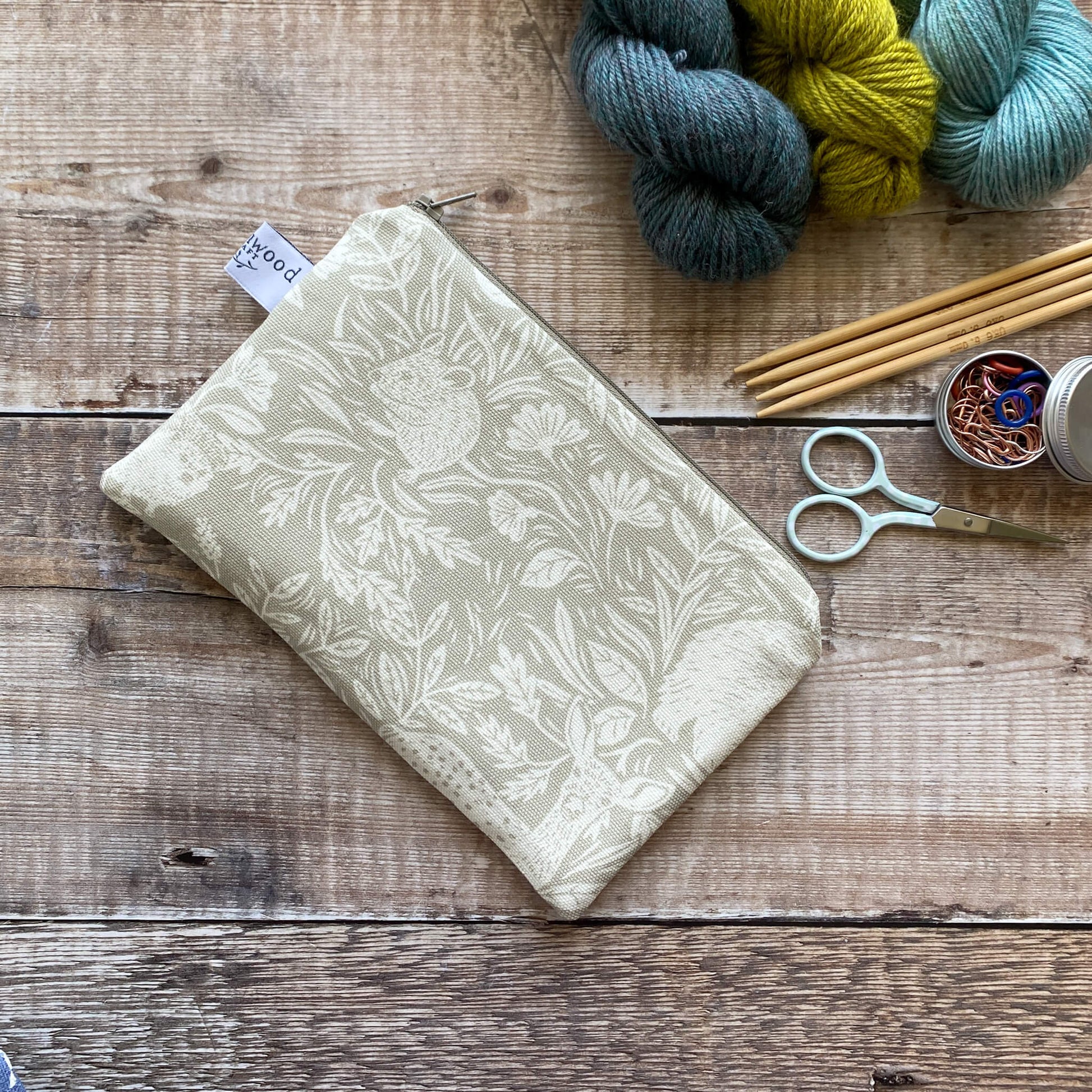 A small zipped pouch hand made using linen coloured countryside themed fabric lies on a wooden table. The pouch is designed to be used to hold knitting and crochet accessories. Next to the pouch are some scissors, knitting needles and stitch markers. 