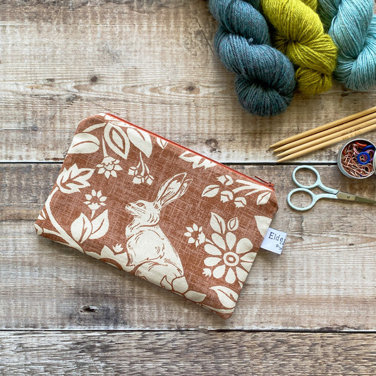 A small zipped pouch hand made using copper coloured heathland themed fabric lies on a wooden table. The pouch is designed to be used to hold knitting and crochet accessories. Next to the pouch are some scissors, knitting needles and stitch markers. 