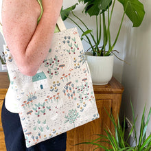 Load image into Gallery viewer, A tote bag is worn on the shoulder of a woman wearing a green top and black trousers. The woman is standing next to green leafy plants and a wooden cabinet. 