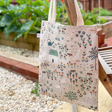 Load image into Gallery viewer, A tote bag hangs from the back of a garden chair in front of a vegetable patch full of luscious green veg. The bag fabric shows a hand drawn cottage garden scene including images of ducks and chickens, vegetables and flowers and a pretty cottage. 