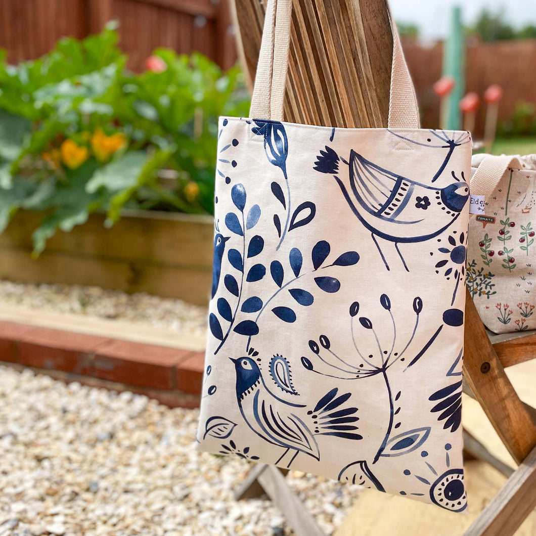 A knitting tote bag hangs gracefully over the back of a garden chair. The tote bag is spacious and made of durable fabric, designed specifically for carrying knitting supplies and projects. The chair is situated in a picturesque outdoor setting, with a lush vegetable patch visible in the background. 