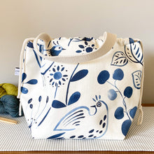 Load image into Gallery viewer,  A knitting project bag placed on a table. The handles are falling down. The bag is made of fabric with a folk art image printed in indigo color. The print depicts various traditional motifs, such as flowers, birds, and geometric patterns. Behind the bag, there are three skeins of yarn, each in different colors. 