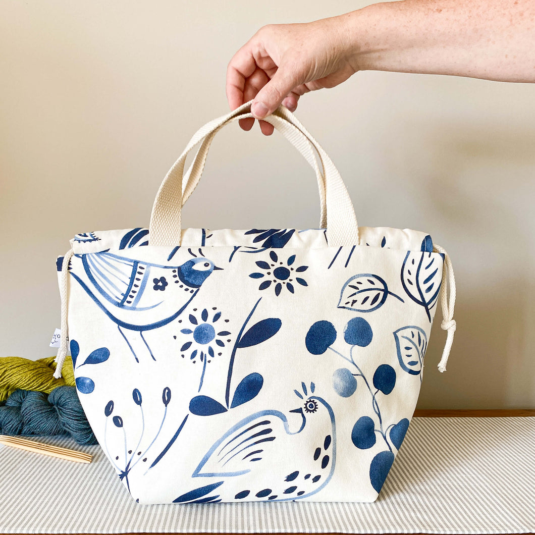  A knitting project bag placed on a table. A hand holds the handles upright. The bag is made of fabric with a folk art image printed in indigo color. Behind the bag, there are three skeins of yarn, each in different colors. 