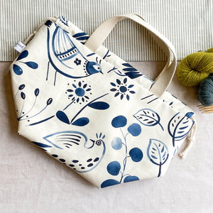 A knitting project bag made out of a Scandi inspired folk art print lies flat on a table. Peeking into the image are two skeins of yarn, one green and one blue, alongside some wooden knitting needles. 