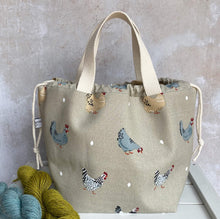 Load image into Gallery viewer, An extra large knitting project bag sits on a bench next to some yarn. The bag fabric features lots of chickens and their eggs. The chickens all look rather cheeky, some are pecking at the ground. The bag has a drawstring closure that is pulled closed and sturdy carry handles. 