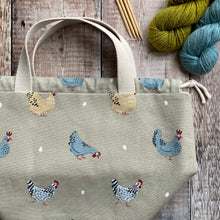 Load image into Gallery viewer, A large chicken project bag lies on a wooden table next to yarn and knitting needles. The bag is made from a fun chicken print and each chicken appears to have laid an egg. 