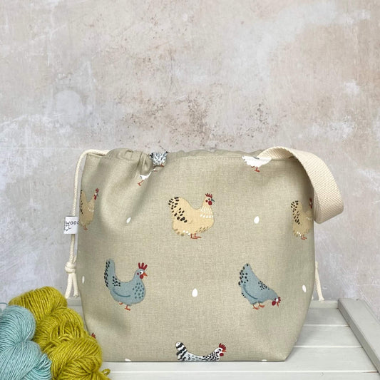 A medium sized hand made knitting project bag sits on a bench next to some yarn. The bag's fabric features cheeky chickens and their eggs on a linen coloured background. 