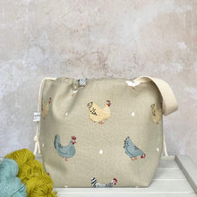 Load image into Gallery viewer, A medium sized hand made knitting project bag sits on a bench next to some yarn. The bag&#39;s fabric features cheeky chickens and their eggs on a linen coloured background. 