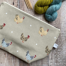 Load image into Gallery viewer, A zippered pouch intended to be used for knitting and crochet projects lies on a wooden table next to two skeins of yarn and some knitting needles. The project bag is covered in hand drawn cheeky chickens. 
