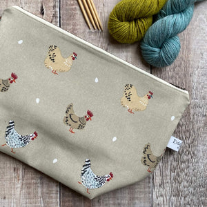 A zippered pouch intended to be used for knitting and crochet projects lies on a wooden table next to two skeins of yarn and some knitting needles. The project bag is covered in hand drawn cheeky chickens. 
