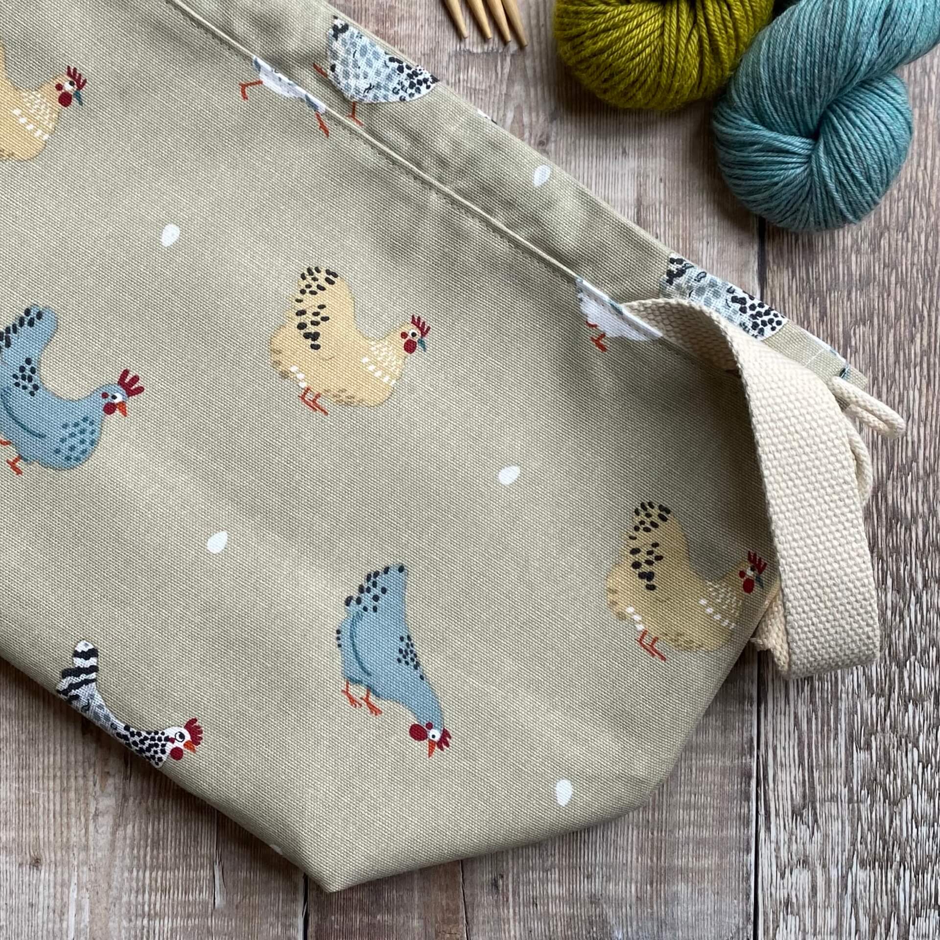 A medium sized knitting project bag lies on a wooden table next to some yarn. It has a sturdy natural coloured handle and images of chickens and their eggs which have been hand drawn. 