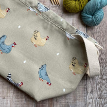 Load image into Gallery viewer, A medium sized knitting project bag lies on a wooden table next to some yarn. It has a sturdy natural coloured handle and images of chickens and their eggs which have been hand drawn. 