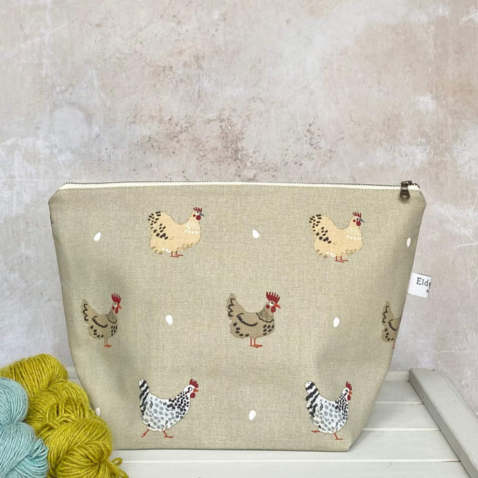 A zipped knitting project bag suitable for medium sized wips sits on a bench next to two skeins of yarn. The bag is hand made using fabric covered in cheeky hand drawn chickens and their eggs. 