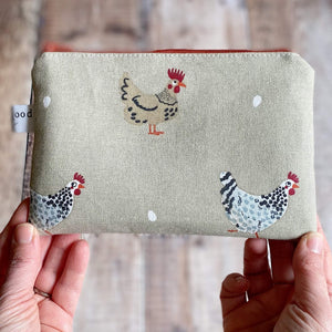 A pair of hands holds up a knitting notions pouch made from chicken fabric. The background is a wooden table .