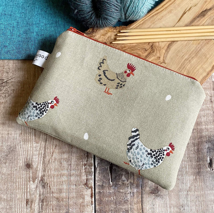 A small zippered pouch lies on a wooden table next to some yarn and knitting needles. The pouch is perfect for keeping knitting notions in and is made from a chicken print. Three chickens can be seen and each of them appears to have laid an egg. 