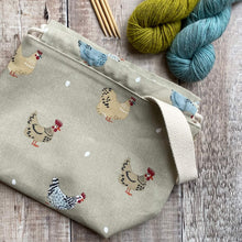 Load image into Gallery viewer, A project bag for knitting featuring cheeky chickens and their eggs lies on a wooden table next to two skeins of yarn and some knitting needles