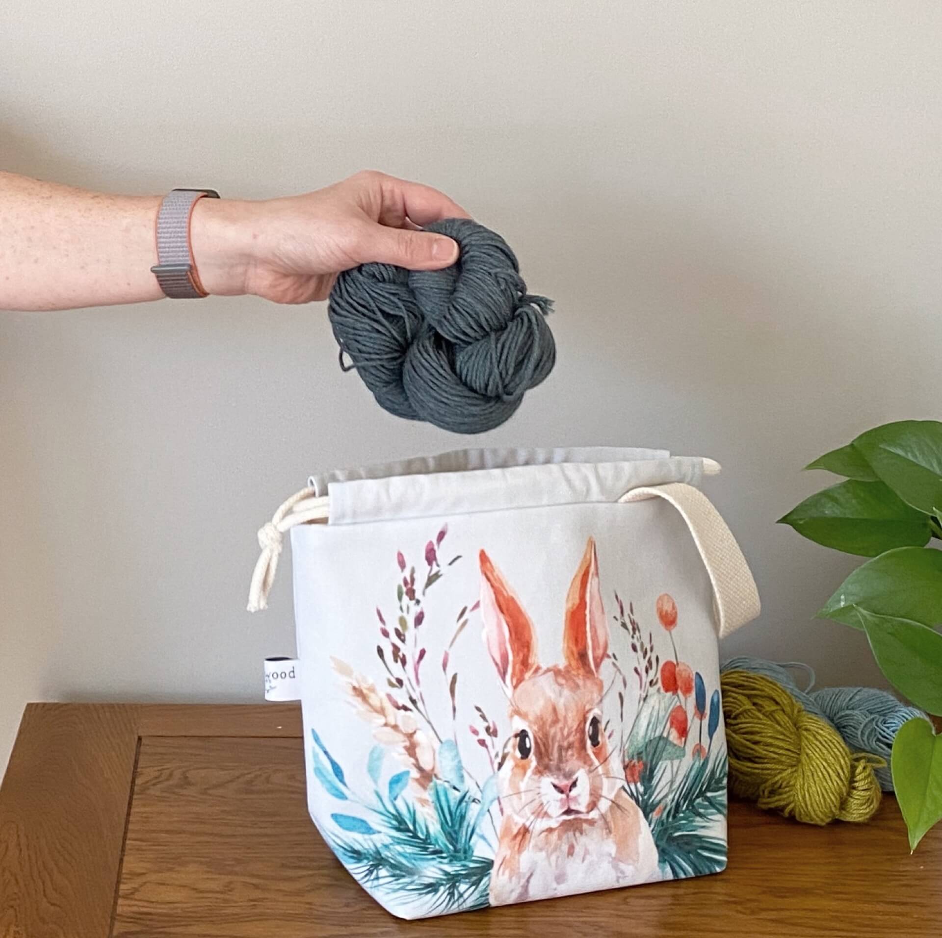 A handmade project bag from Eldenwood Craft is sitting a wooden table next to two skeins of yarn and a plant. The project bag features a watercolour hare surrounded by leaves and shoots and a skein of blue yarn is being placed into the bag. 