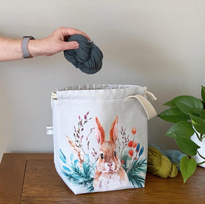 A handmade project bag from Eldenwood Craft is sitting a wooden table next to two skeins of yarn and a plant. The project bag features a watercolour hare surrounded by leaves and shoots and a skein of blue yarn is being placed into the bag. 