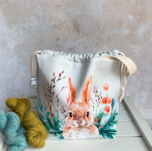 Load image into Gallery viewer, A knitting project bag handmade by Eldenwood Craft featuring a watercolour hare print. The project bag is sitting on a bench next to two skeins of yarn. 