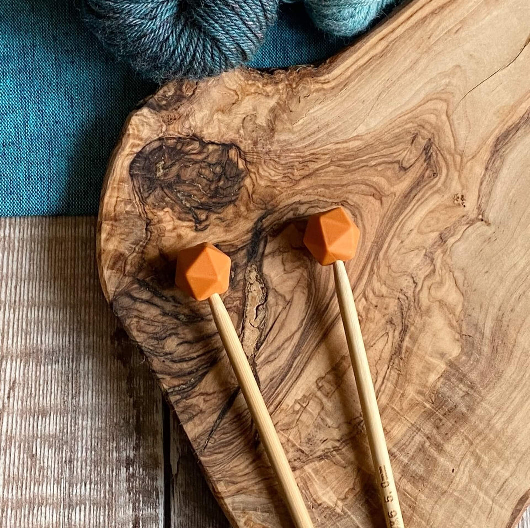 Burnt Orange needle protectors to stop stitches escaping from knitting needles. A pair sit on a wooden table attached to knitting needles. 