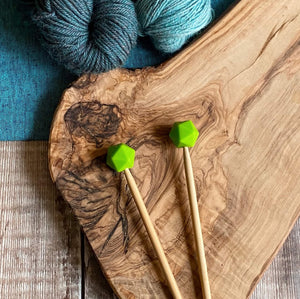 Chartreuse needle protectors to stop stitches escaping from knitting needles. A pair sit on a wooden table attached to knitting needles. 