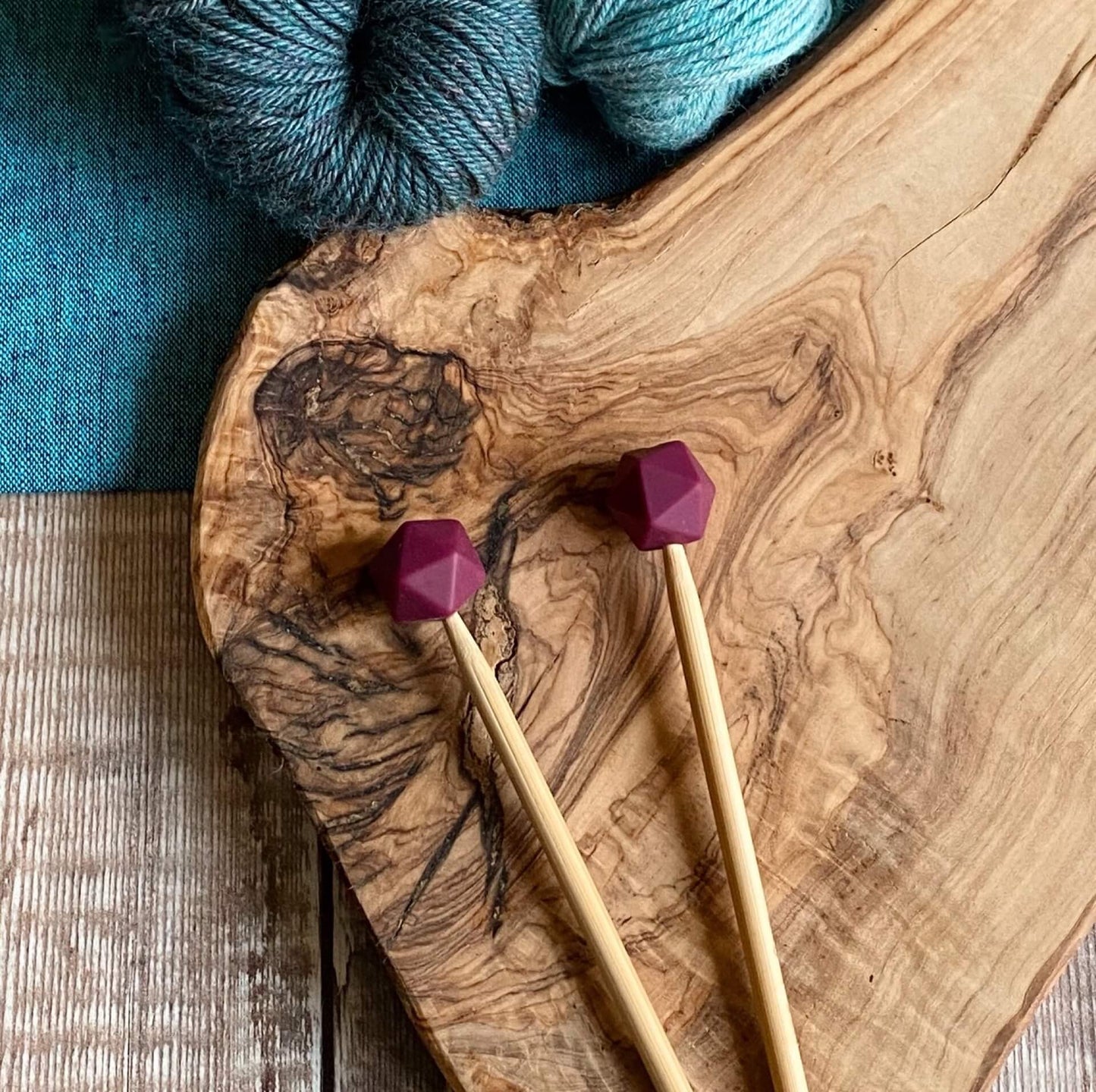 Plum needle protectors to stop stitches escaping from knitting needles. A pair sit on a wooden table attached to knitting needles. 