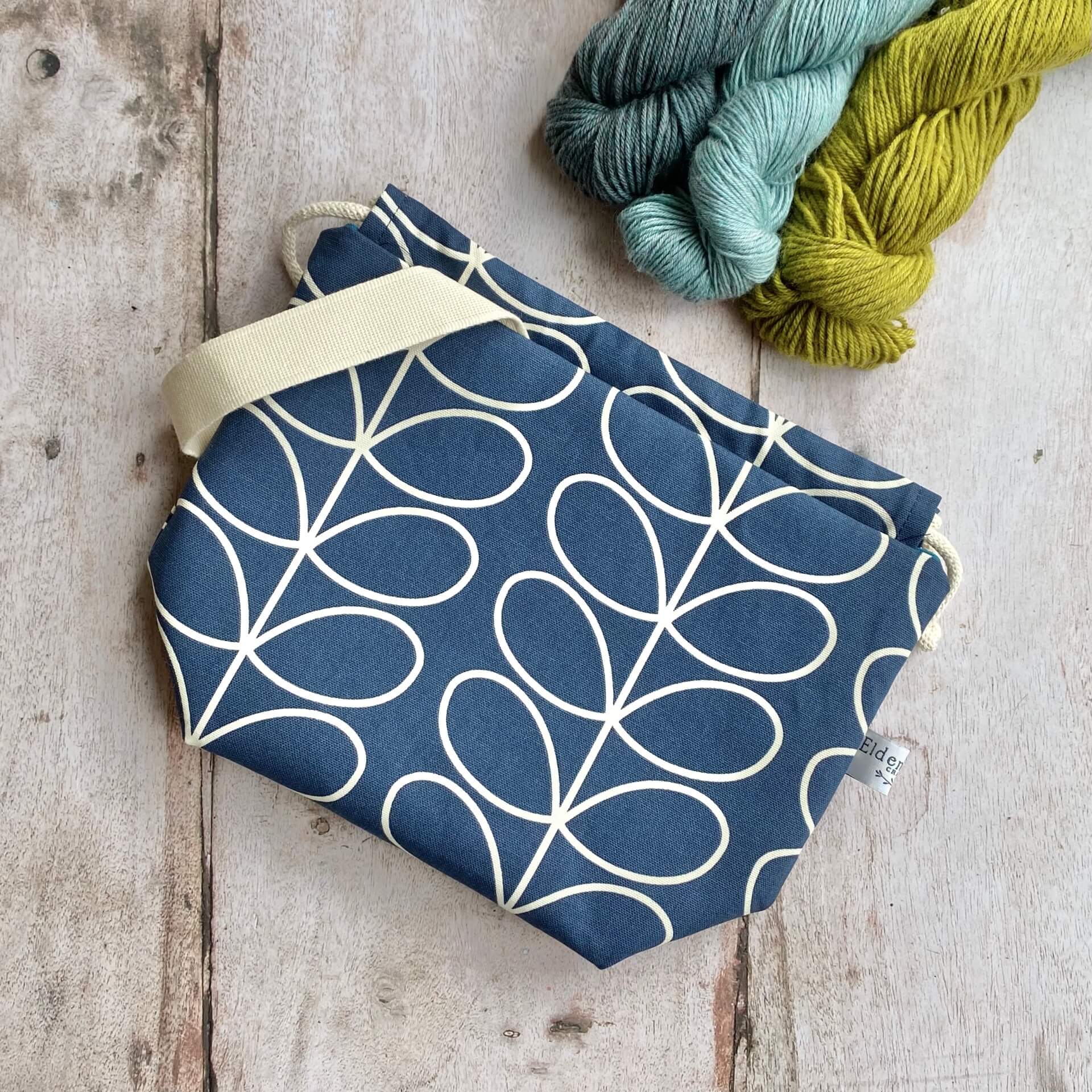 A handmade knitting project bag from Eldenwood Craft lies on a wooden table next to some yarn. The bag is made from navy fabric in a classic Orla Kiely linear stem print.
