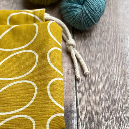 A knitting project bag shown in close up. The bag lies on a wooden table next to two skeins of yarn. 