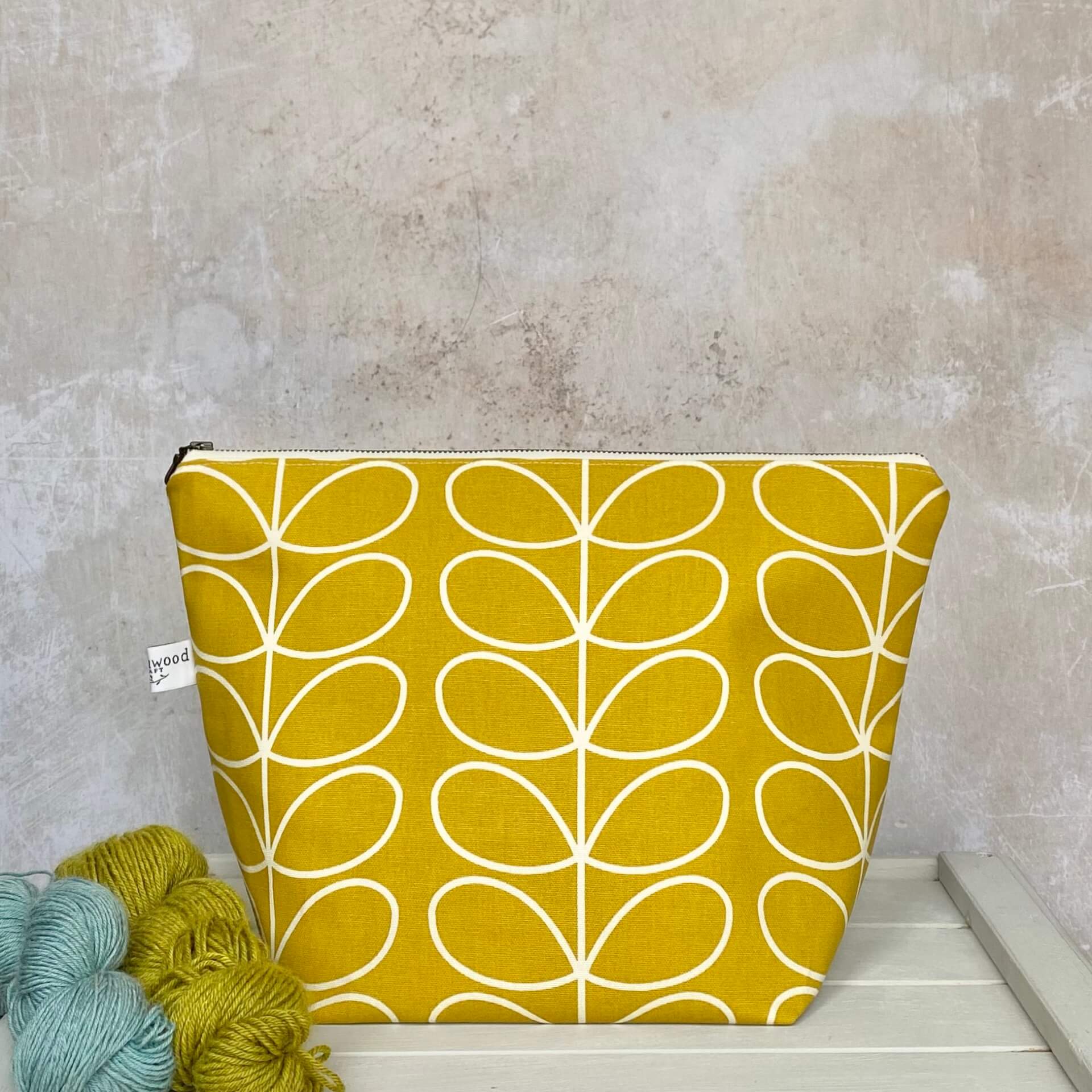 A zippered pouch sits on a bench next to some yarn. The bag is handmade from a cheerful sunshine yellow Orla Kiely fabric and is intended for use as a knitting project bag. 