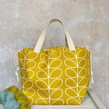 Load image into Gallery viewer, A large knitting project bag in a sunshine yellow Orla Kiely print sits on a bench next to two skeins of yarn. The project bag stands upright and its drawstrings are pulled closed. 