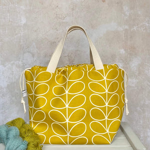 A large knitting project bag in a sunshine yellow Orla Kiely print sits on a bench next to two skeins of yarn. The project bag stands upright and its drawstrings are pulled closed. 