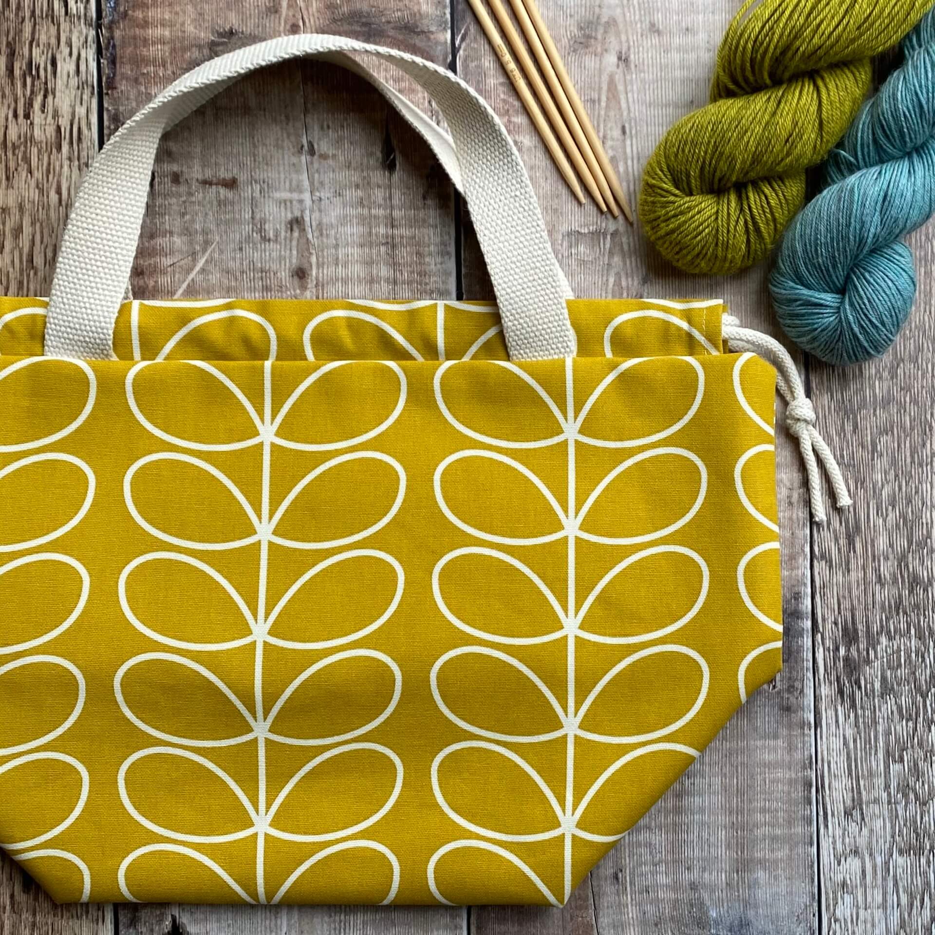 A large knitting project bag lies on a wooden table next to yarn and knitting needles. The bag is handmade using fabric in a cheerful yellow Orla Kiely print. The bag is part of the Eldenwood Craft collection of project bags for knitting. 