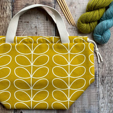 Load image into Gallery viewer, A large knitting project bag lies on a wooden table next to yarn and knitting needles. The bag is handmade using fabric in a cheerful yellow Orla Kiely print. The bag is part of the Eldenwood Craft collection of project bags for knitting. 