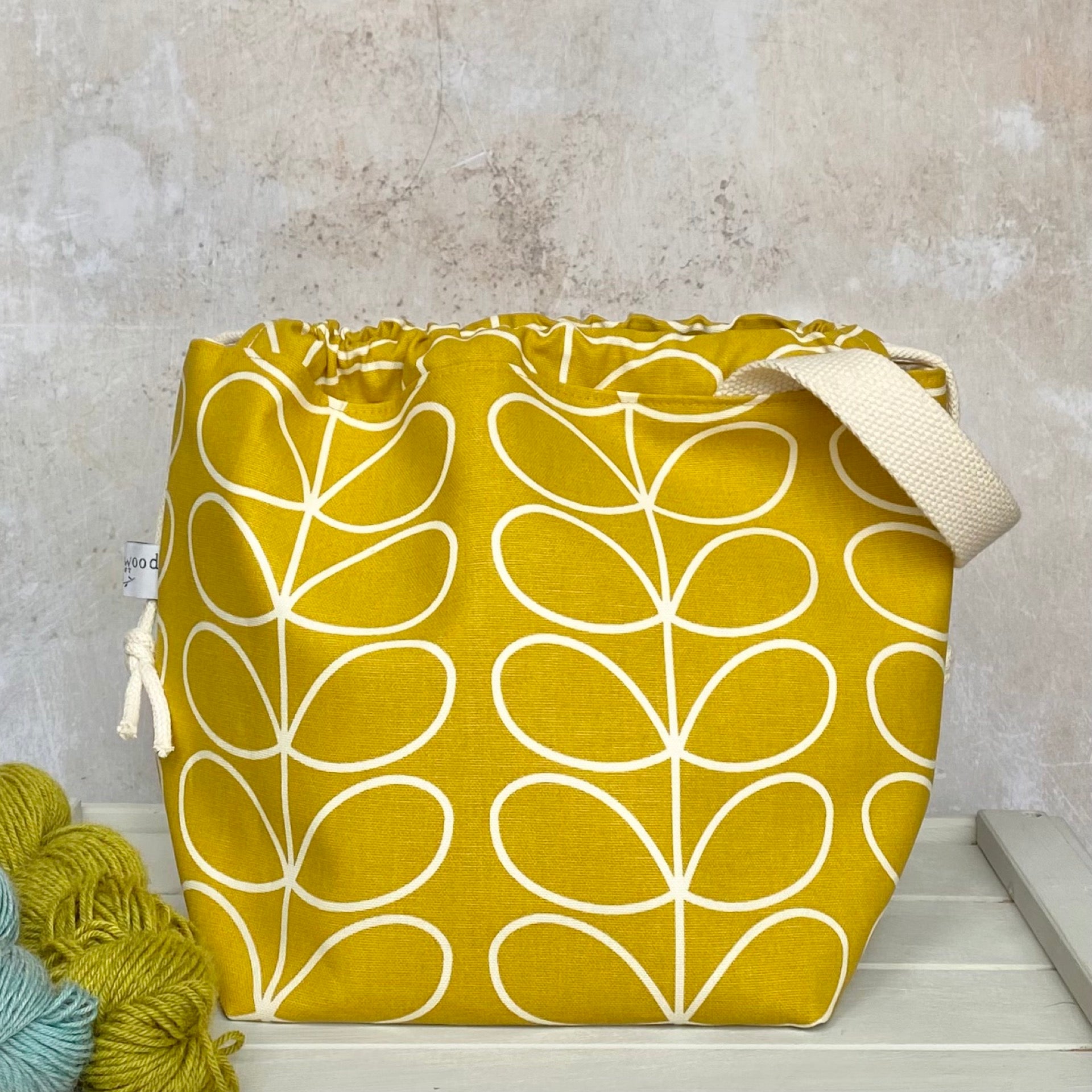 A handmade project bag sits on a bench next to two skeins of yarn. The bag is handmade using a sunshine yellow Orla Kiely fabric. 